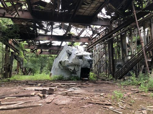 An abandoned building with a collapsed roof, overgrown with plants and debris scattered on the ground.