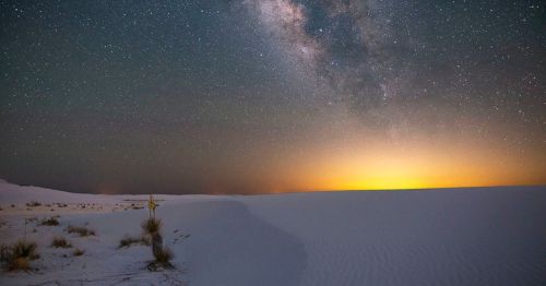 A serene desert landscape under a starry sky, with the Milky Way and a glowing horizon at dusk.