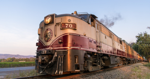 A vintage train with a maroon and silver exterior travels along a scenic track during sunset.