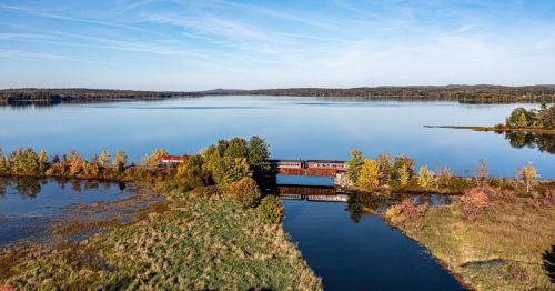A serene landscape featuring a calm lake, autumn foliage, and a train crossing a bridge over water.