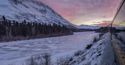 A scenic view of a snowy landscape with mountains, a frozen river, and a train reflecting the sunset colors.