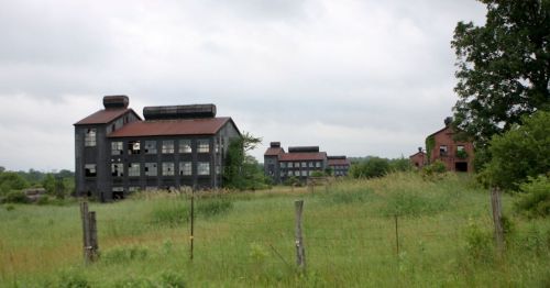Abandoned industrial buildings with red roofs, surrounded by overgrown grass and trees under a cloudy sky.