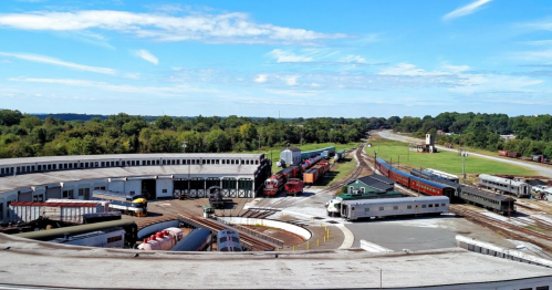 A panoramic view of a train yard with multiple trains and tracks under a clear blue sky. Green trees in the background.