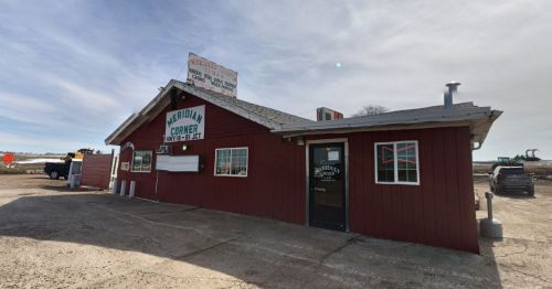 A red wooden building with a sign reading "Meridian Corner" and large windows, set against a cloudy sky.