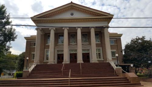 A large, classical-style building with columns and stairs, set against a cloudy sky.