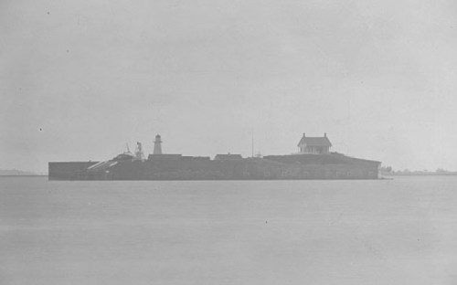 A historic black-and-white image of a coastal fort with a lighthouse and a building on a small island.