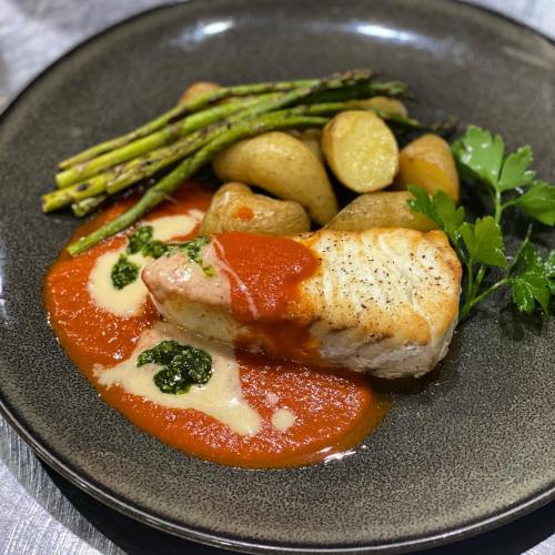 A plate featuring grilled fish with tomato sauce, asparagus, baby potatoes, and a drizzle of green sauce.