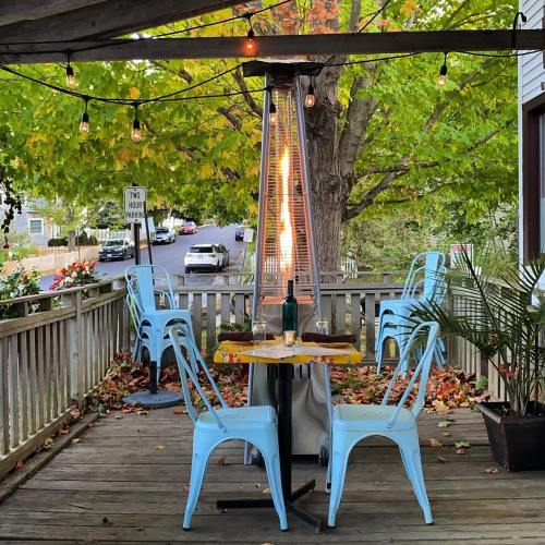 A cozy outdoor dining area with blue chairs, a table, and a patio heater surrounded by autumn leaves and trees.