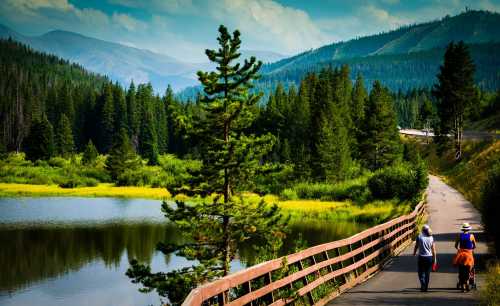 A scenic path by a lake, with two people walking and lush green trees against a backdrop of mountains and blue sky.