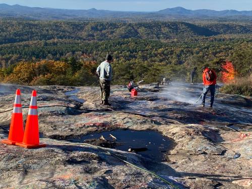 Workers clean a rocky surface with spray equipment, surrounded by autumn foliage and distant mountains. Traffic cones are visible.