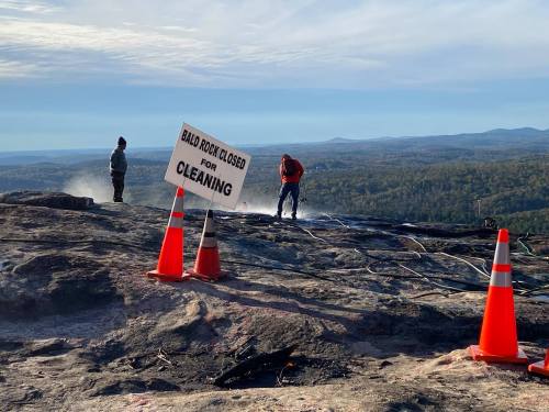 Two workers clean a rocky area with a "Bald Rock Closed for Cleaning" sign and traffic cones in the foreground.