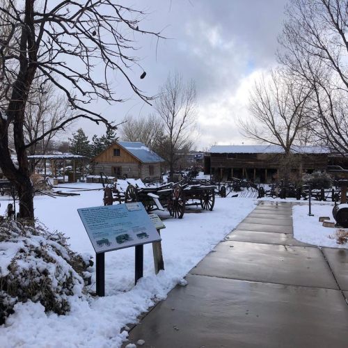 A snowy outdoor museum with historical buildings and artifacts, featuring a pathway and informational sign.