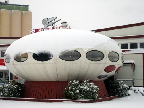 A futuristic, UFO-shaped building covered in snow, surrounded by greenery and located near a commercial area.