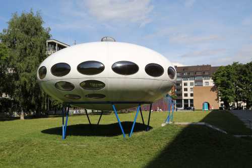 A futuristic, white UFO-shaped structure with large windows, sitting on a grassy area near buildings.