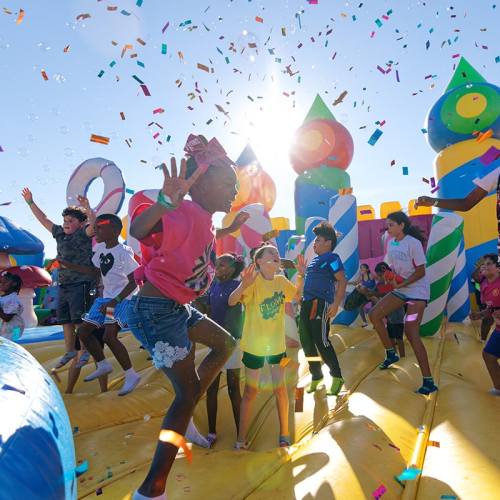 Children joyfully jumping on a bouncy castle, surrounded by colorful confetti and bubbles under a bright sky.