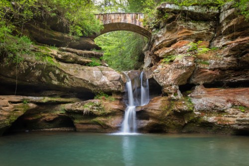 A serene waterfall cascades into a clear pool, framed by rocky cliffs and a rustic stone bridge above.