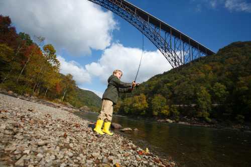 A child in yellow boots fishes by a river under a large bridge, surrounded by trees and a blue sky.