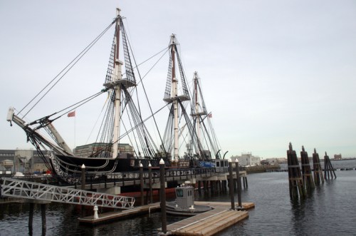 A historic ship docked at a harbor, with masts and rigging visible against a cloudy sky.
