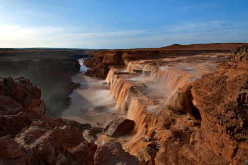 A stunning waterfall cascades over rocky cliffs into a river, surrounded by rugged terrain and a clear blue sky.
