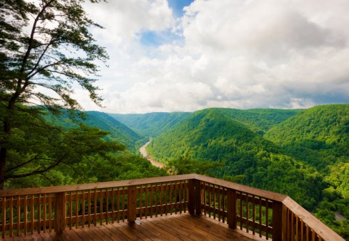 A scenic view from a wooden deck overlooking lush green mountains and a winding river under a partly cloudy sky.