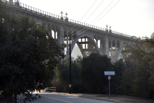 A large, arched bridge rises above a tree-lined road, with power lines running alongside it in a misty atmosphere.