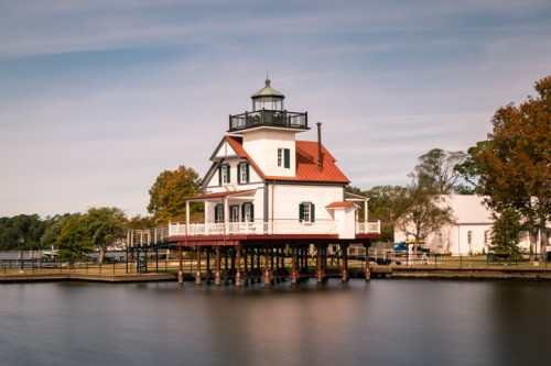 A charming lighthouse on stilts by a calm lake, surrounded by trees and a clear blue sky.