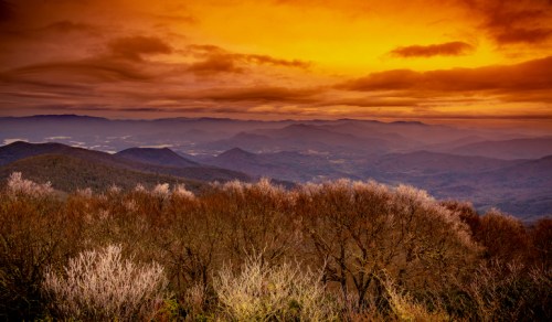 A stunning sunset over layered mountains, with vibrant orange skies and silhouetted trees in the foreground.