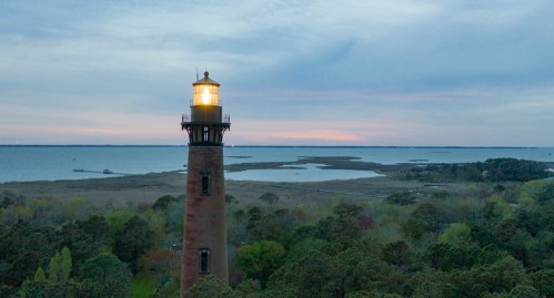 A tall lighthouse stands by a calm body of water, surrounded by trees, under a pastel sky at dusk.