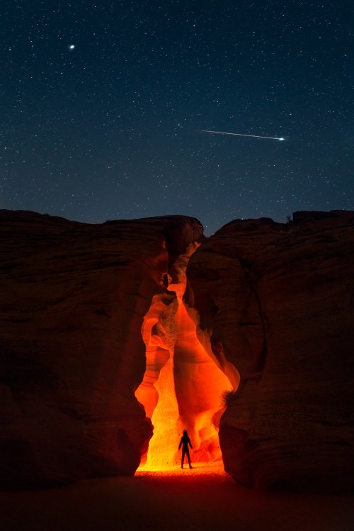A silhouette stands in a glowing canyon under a starry sky, with a shooting star above.