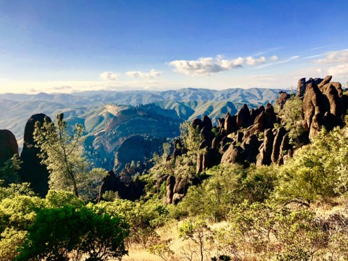 A panoramic view of rugged mountains and rocky formations under a clear blue sky, surrounded by lush greenery.