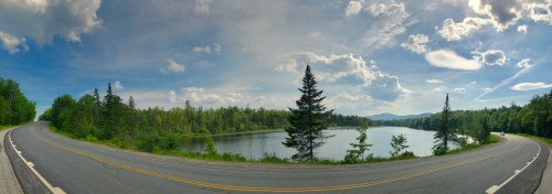A scenic view of a winding road beside a calm lake, surrounded by lush greenery and a blue sky with fluffy clouds.