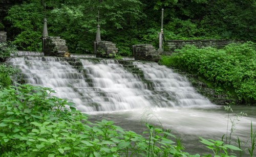 A serene waterfall cascades over stone steps, surrounded by lush greenery and trees.