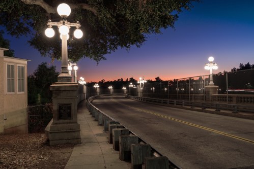A quiet, illuminated bridge at dusk, lined with vintage street lamps and a colorful sunset in the background.