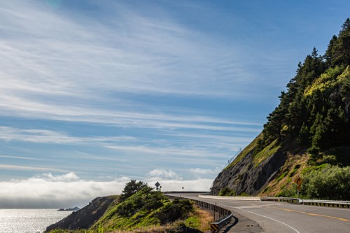 A winding coastal road curves along a cliffside, with lush greenery and the ocean in the background under a blue sky.