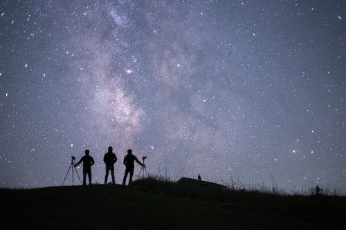 Three silhouetted figures stand on a hill, gazing at a starry sky filled with the Milky Way and distant stars.