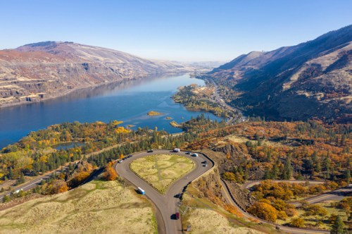 Aerial view of a winding road with cars, overlooking a river and autumn-colored hills. Clear blue sky above.