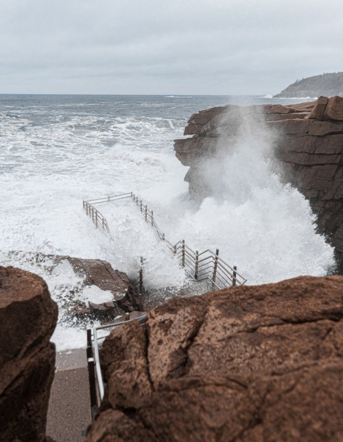 Waves crash against rocky cliffs near a staircase leading down to the ocean on a cloudy day.