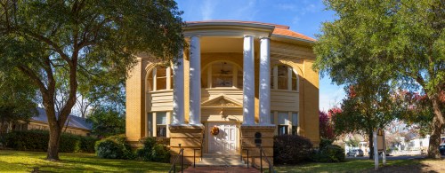 A historic building with large columns, a red roof, and lush greenery surrounding it on a sunny day.