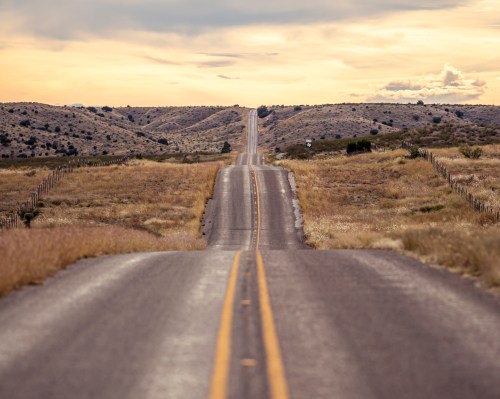 A winding road stretches through a grassy landscape, leading towards distant hills under a cloudy sky.