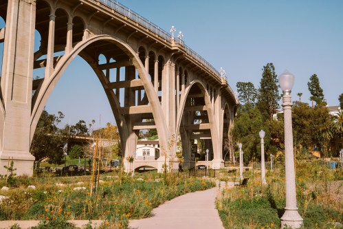 A concrete bridge with arches spans over a park, featuring a pathway and greenery under a clear blue sky.