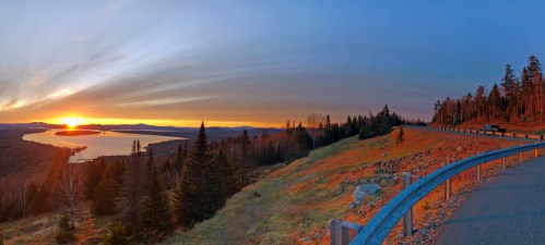 A panoramic view of a sunset over a lake, surrounded by trees and rolling hills, with a road in the foreground.