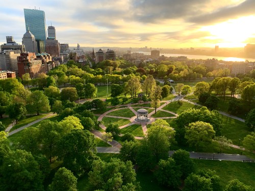 Aerial view of a lush green park with a gazebo, surrounded by city buildings and a sunset sky in the background.