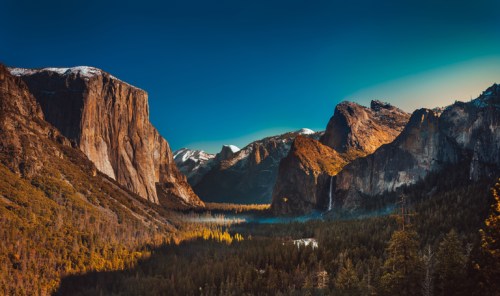 A stunning landscape of mountains and a valley, with a waterfall and vibrant autumn foliage under a clear blue sky.