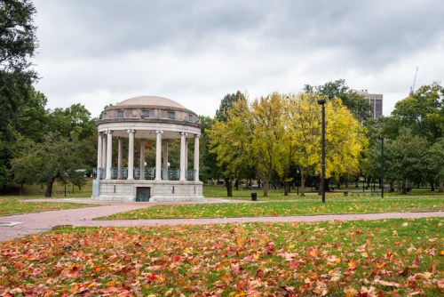 A gazebo surrounded by colorful autumn leaves in a park, with overcast skies and trees in the background.