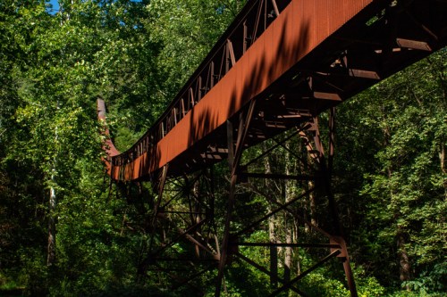 A rusted metal bridge stretches over lush green trees in a forested area.
