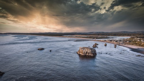A scenic coastal view featuring rocky formations, a sandy beach, and dramatic clouds over the water at sunset.