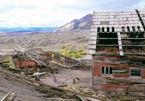 Abandoned, weathered buildings amidst a barren landscape with mountains in the background and scattered debris.