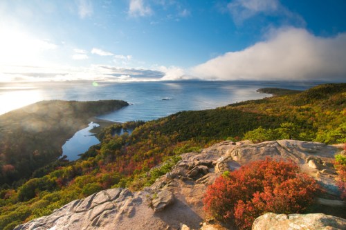 A scenic view of a coastline with vibrant autumn foliage, calm waters, and a cloudy sky in the background.