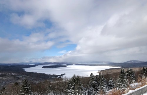 A snowy landscape with a frozen lake, surrounded by trees and mountains under a partly cloudy sky.