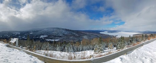 A panoramic winter landscape featuring snow-covered mountains, trees, and a frozen lake under a partly cloudy sky.